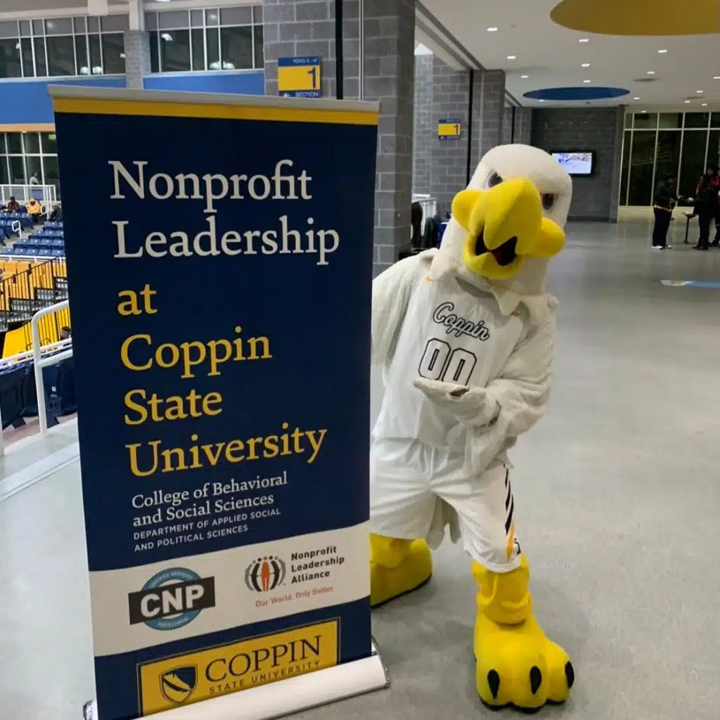 Campus Partner, Coppin State University Mascot of a White Eagle posing next to a Nonprofit Leadership Alliance and Certified Nonprofit Professional Sign at an indoor sporting stadium