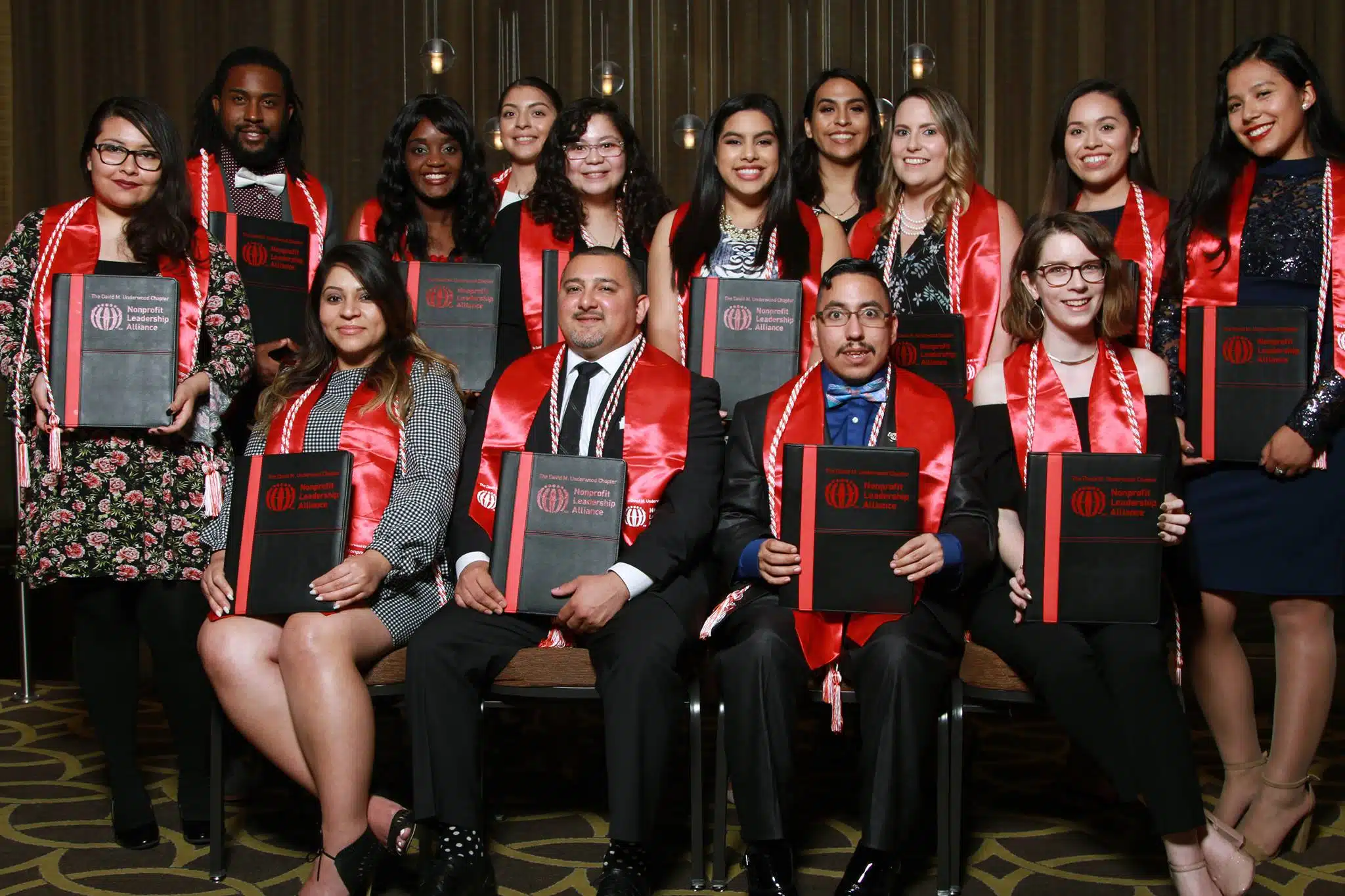 A group of diverse college graduates holding their Certified Nonprofit Professional Certificates in their hands with dark red graduation sashes from campus partner university of houston