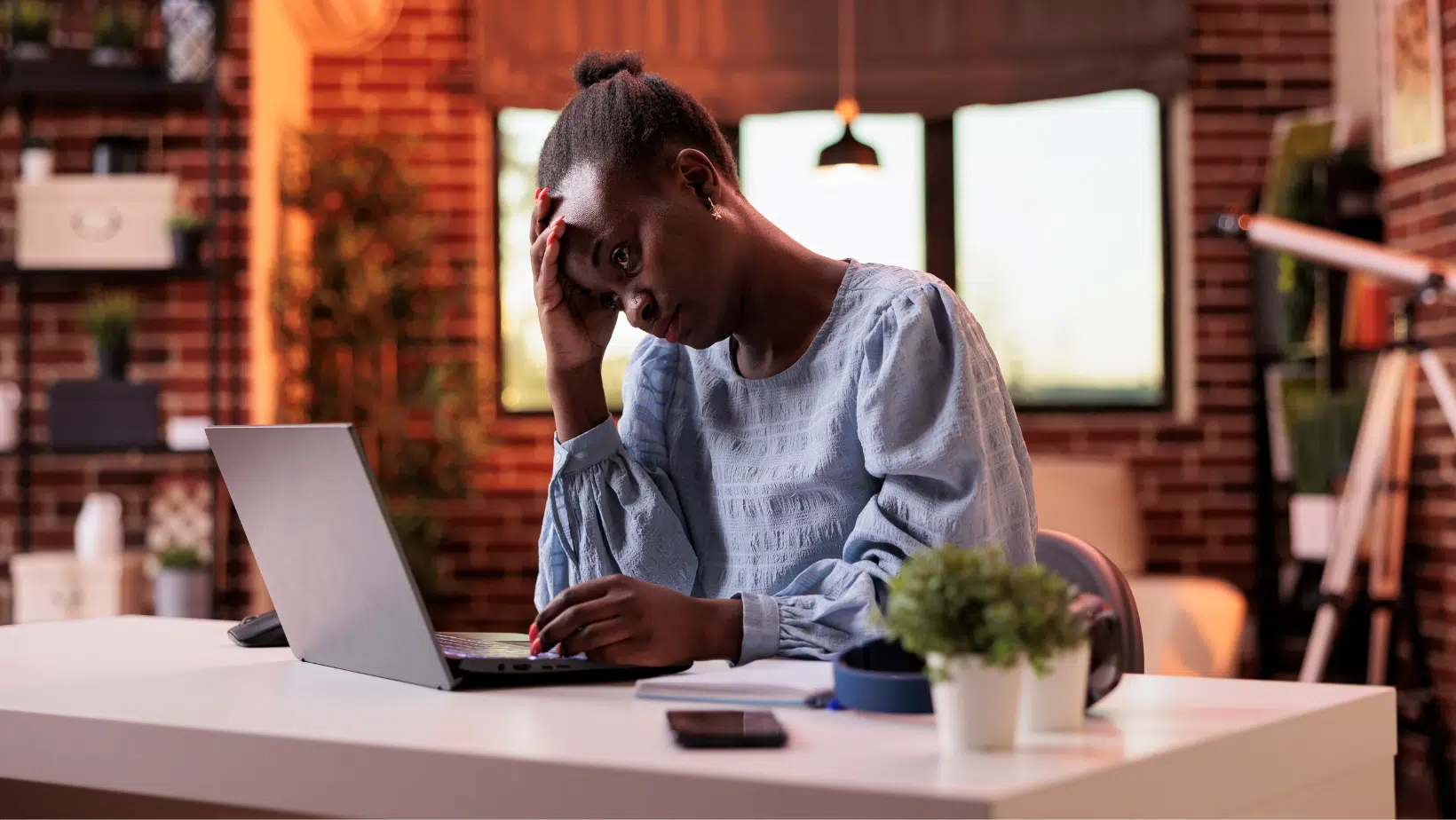 African American Woman sitting at a white desk in a professional office. She has a hand leading on her forehead and is dealing with burnout