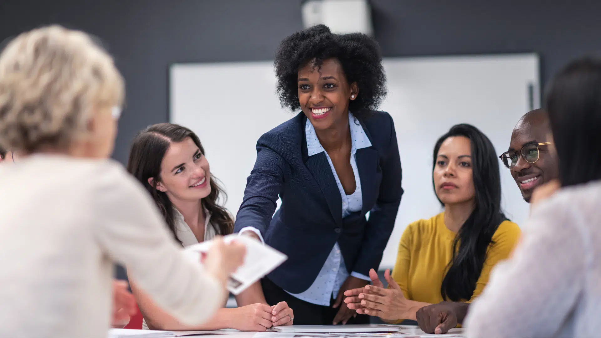 A confident African American woman is meeting with her nonprofit boards members. The woman leading the group is standing at the head of the table smiling while passing a document out.