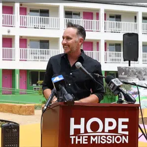 Rowan Vansleve dressed in a black professional button down standing at a wooden podium speaking and addressing a crowd