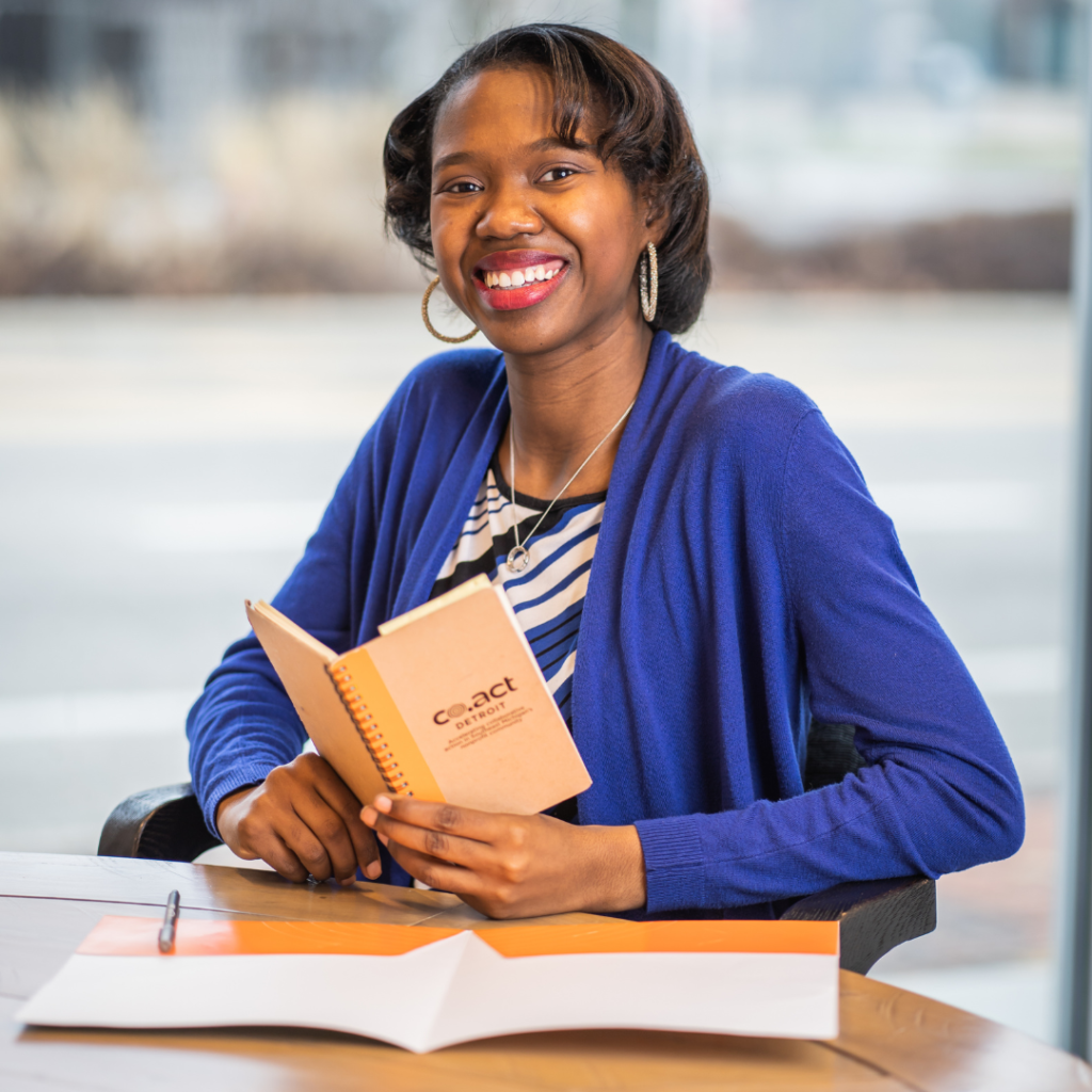 Detriot top nonprofit professional, Charnae Sanders smiling in an office while wearing a black and white blouse and purple cardigan while holding a Co.act Detroit branded notebook