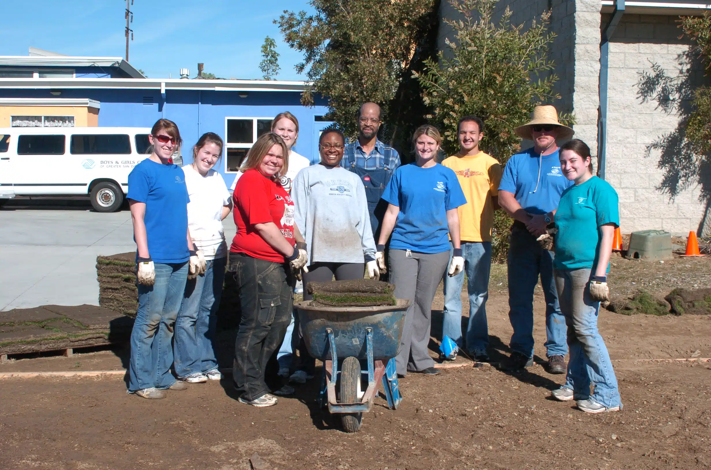 Large group of individuals working outside and volunteering. Once is holding a wheel barrel full of dirt