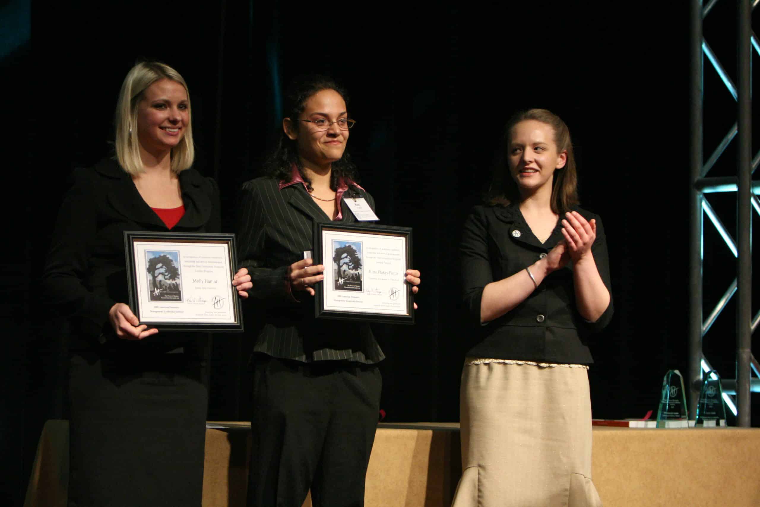 Three women on a stage. Two of them are holding certificates and the third is clapping in celebration