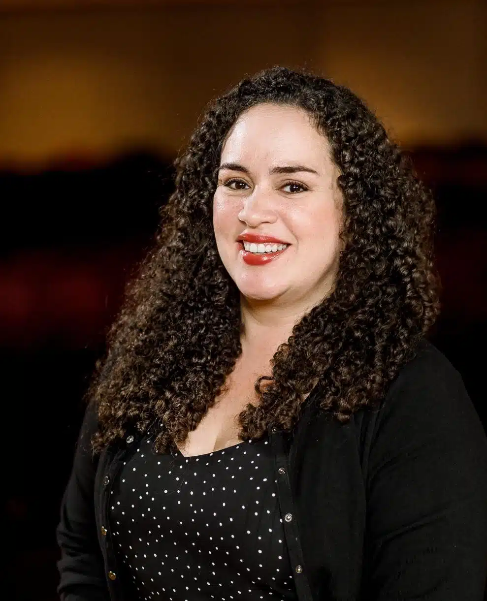 Headshot of a curly-haired brunette woman in a black blazer and black top.