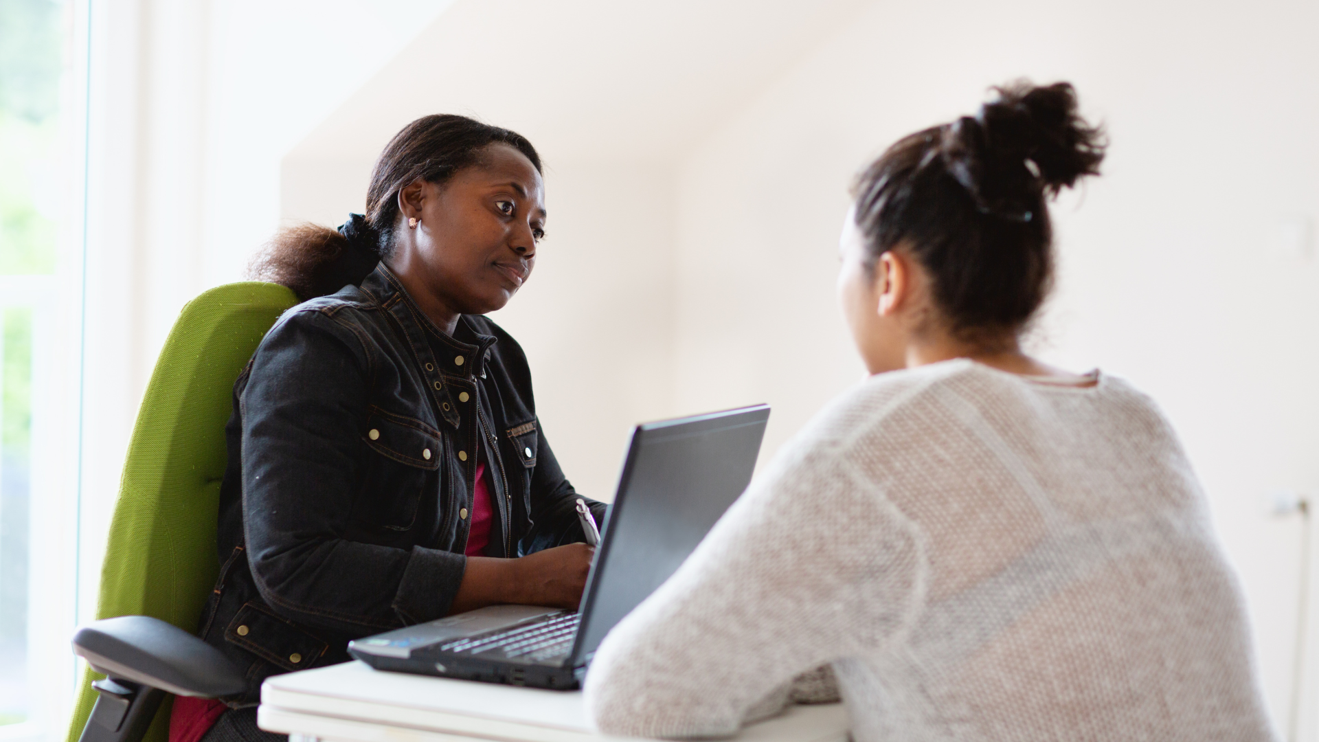 An African American Woman in a denim jacket looks serious and is speaking to a young individual in a white sweater. The woman is on her computer leveraging ai in social work