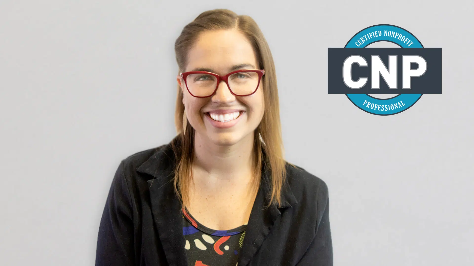 Nicole Olivia Rose, CNP smiling. Caucasian woman with red glasses and professional blazer smiling in front of a grey background