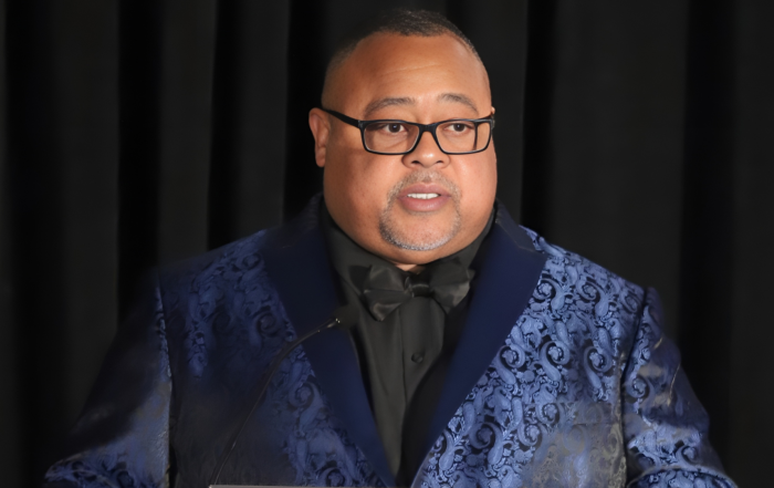 African American Man, Everette Pearsall, stands smiling in a blue and black tuxedo. He is speaking at a podium at an event