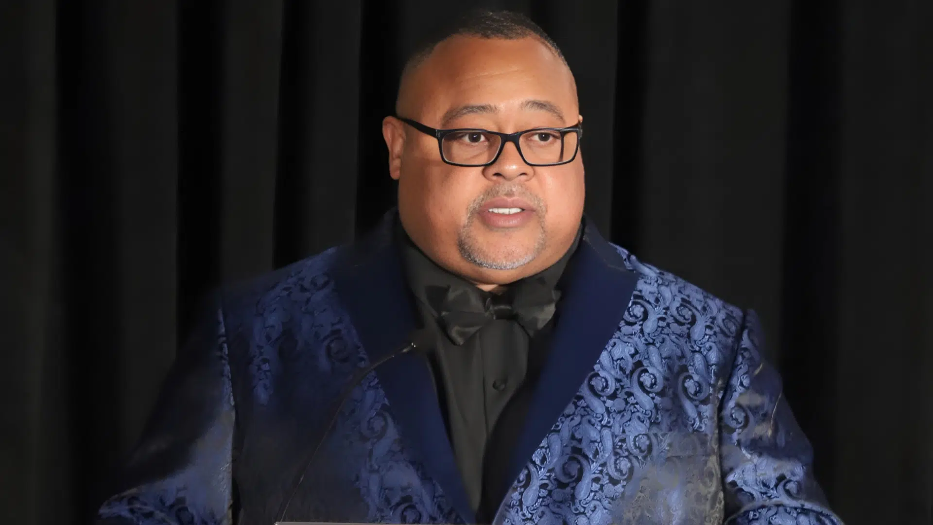 African American Man, Everette Pearsall, stands smiling in a blue and black tuxedo. He is speaking at a podium at an event