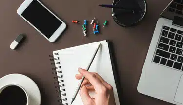 Top view of a desk featuring a laptop, cell phone, push pins, and a hand writing in a notebook drafting a nonprofit's annual report